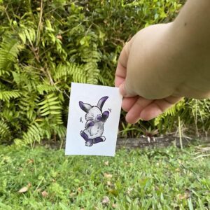 A white person holding a sticker of a purple, gray, and white bunny in front of grass and a garden.