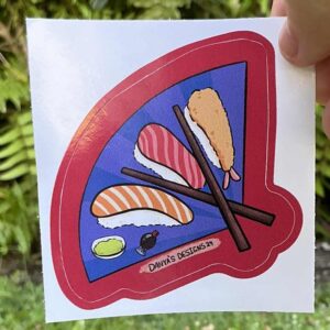 Sticker of salmon, tuna, and prawn sushi where the plate is a paper fan and the chop sticks are the wood that holds the fan together.