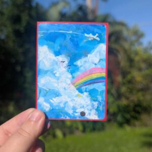 A hand holding up a hand painted artwork of fluffy whight clouds with a rainbow flowing out of them, small birds up and to the lift of the rain bow and a plane in the top right of the artwork all against a blue sky.