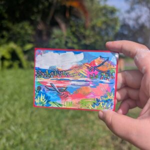 Hand holding up a hand painted artwork of a city across a lake. The artwork uses bright colours through out.
