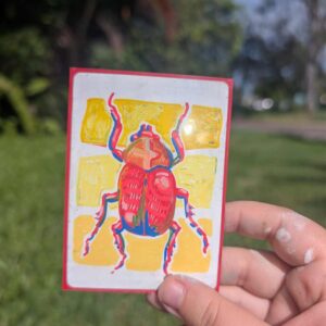 A hand holding up a hand painted artwork of a red beatle with strips of yellow behind the beatle.